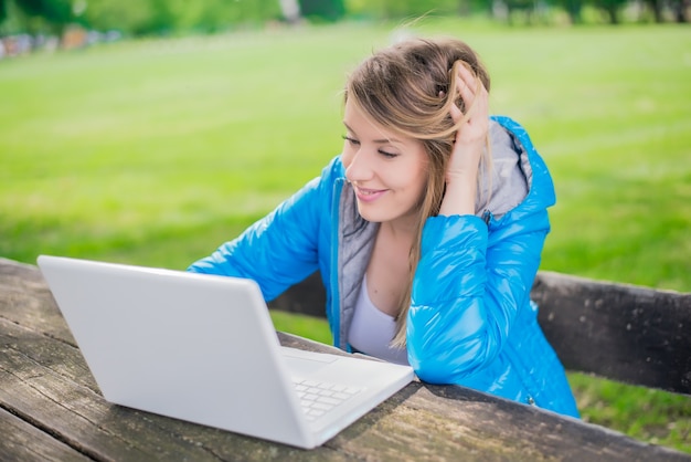 Bella donna studente sta utilizzando un computer portatile e seduto su una panchina nel campus universitario. Donna sorridente sta lavorando su un computer all&#39;aperto nel parco universitario. Studente all&#39;aperto concetto.