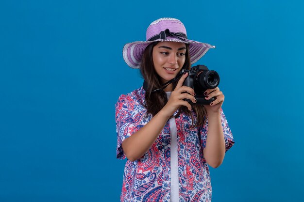 Bella donna turistica in cappello estivo con macchina fotografica con il sorriso sul viso positivo e felice in piedi sopra lo spazio blu isolato