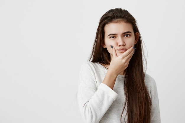 Bella donna vestita casualmente con lunghi capelli lisci scuri che hanno un'espressione dubbiosa, tenendo il mento, cercando di trovare la soluzione migliore. Linguaggio del corpo