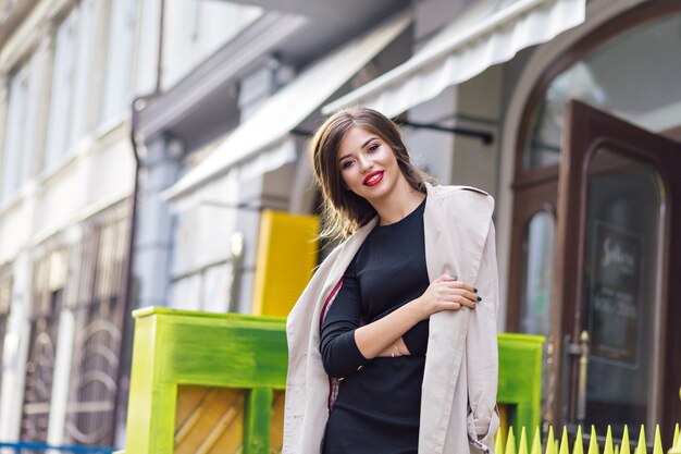 Bella donna vestita in abito nero e trench beige con acconciatura alla moda e labbra rosse in strada