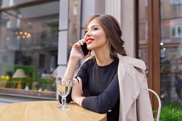 Bella donna vestita in abito nero e trench beige con acconciatura alla moda e labbra rosse su una terrazza, parlando al telefono