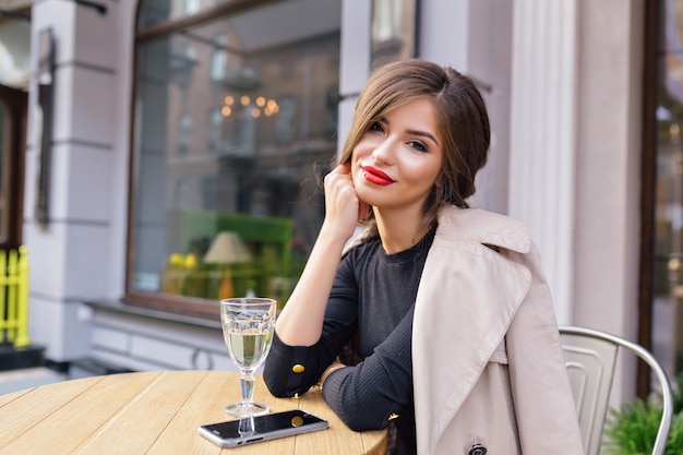 Bella donna vestita in abito nero e trench beige con acconciatura alla moda e labbra rosse su una terrazza