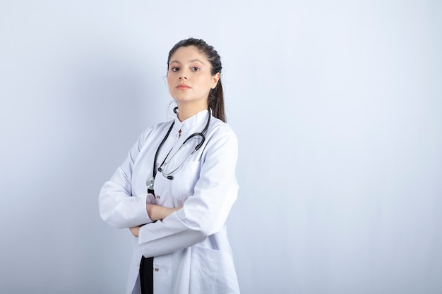 Bella dottoressa in camice bianco in piedi con le braccia incrociate sul muro bianco.