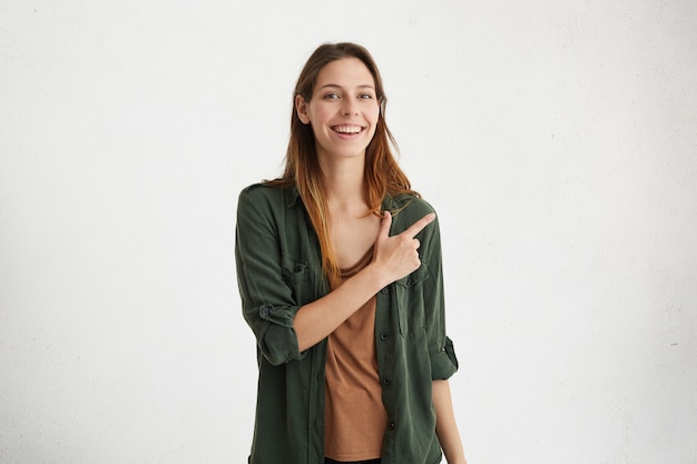 bella e allegra giovane femmina che indossa abiti casual tenendo il dito indice puntato verso il muro bianco bianco con copyspace