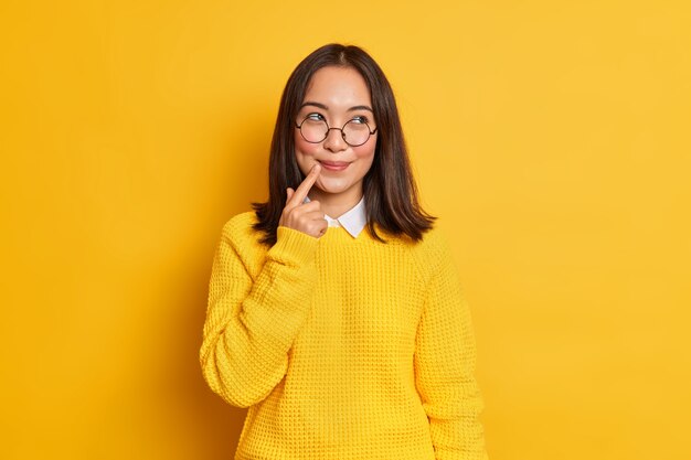 Bella e sognante giovane donna asiatica con i capelli scuri tiene il dito vicino alle labbra indossa un maglione e occhiali trasparenti rotondi.
