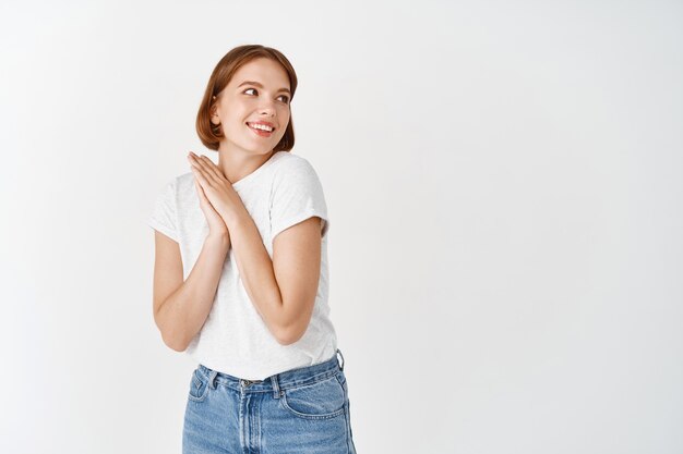 Bella e tenera ragazza naturale, che guarda lontano con le mani premute insieme, guardando qualcosa di adorabile, in piedi sul muro bianco