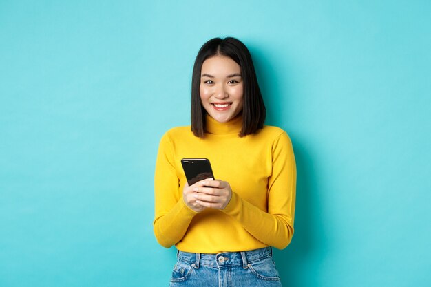 Bella ed elegante donna asiatica che fa shopping online sul telefono cellulare, in piedi su sfondo blu