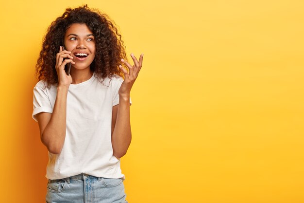 Bella femmina allegra con capelli afro ricci si blocca sul telefono con il migliore amico durante il tempo libero