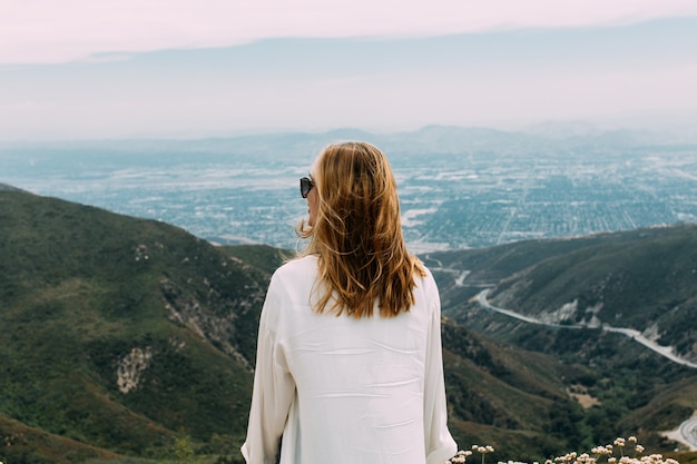 Bella femmina bionda con occhiali da sole e una camicia bianca in piedi sulla cima di una collina nella natura