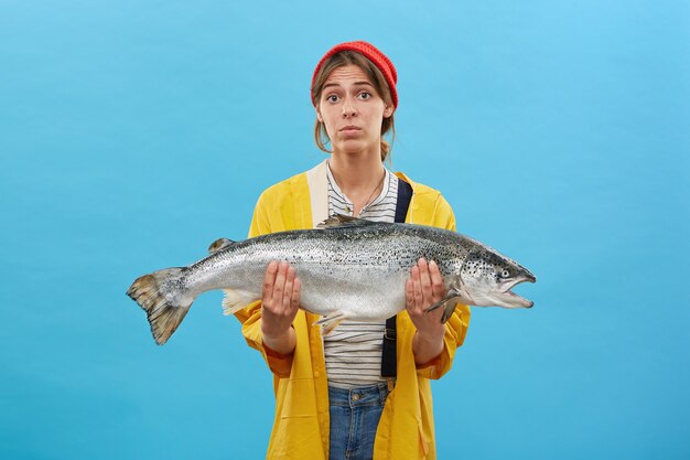 bella femmina che indossa cappello rosso, impermeabile giallo e che tiene in mano un enorme pesce