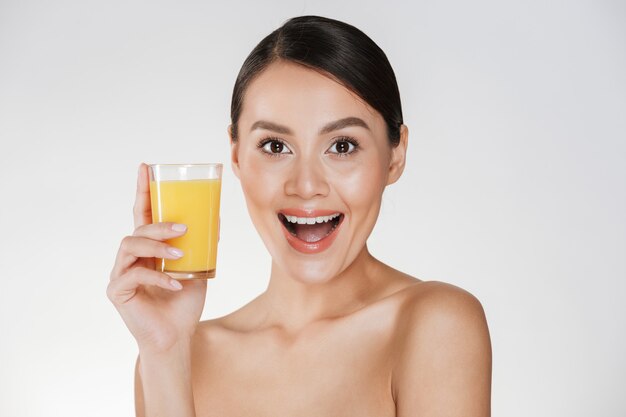 Bella foto di donna seminuda con i capelli scuri in panino e ampio sorriso che beve il succo d'arancia da un vetro trasparente, isolato su un muro bianco