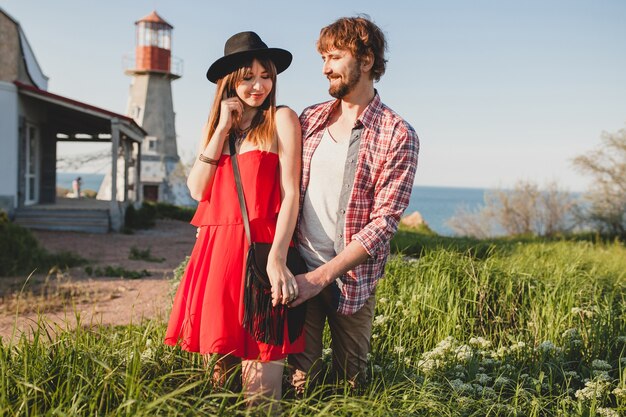 Bella giovane coppia elegante innamorata in campagna, stile bohémien indie hipster, vacanza del fine settimana, vestito estivo, vestito rosso, erba verde, tenendosi per mano, sorridente