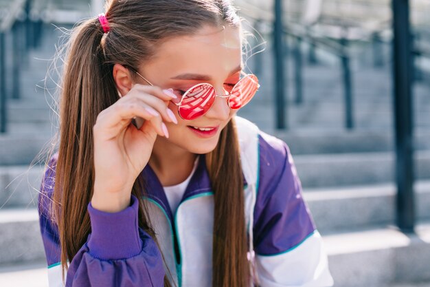 Bella giovane donna alla moda in vestiti alla moda indossando occhiali da sole rosa e sorridente