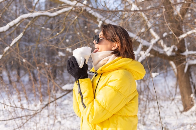 Bella giovane donna allegra in una foresta di inverno paesaggio innevato in occhiali da sole con una tazza piena di neve divertendosi