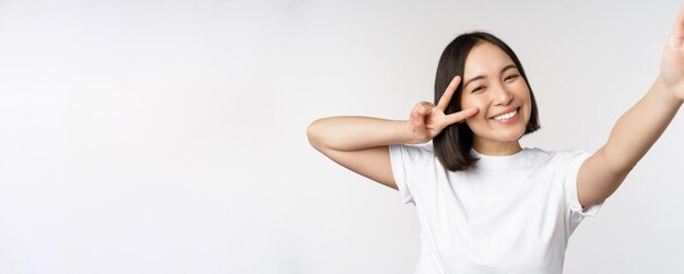 Bella giovane donna asiatica che prende selfie in posa con pace vsign sorridente felice scattare foto in posa su sfondo bianco