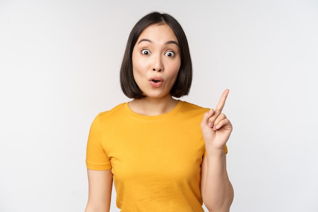 Bella giovane donna asiatica che punta il dito verso l'alto sorridendo e guardando divertito la fotocamera che mostra l'annuncio pubblicitario su sfondo bianco superiore