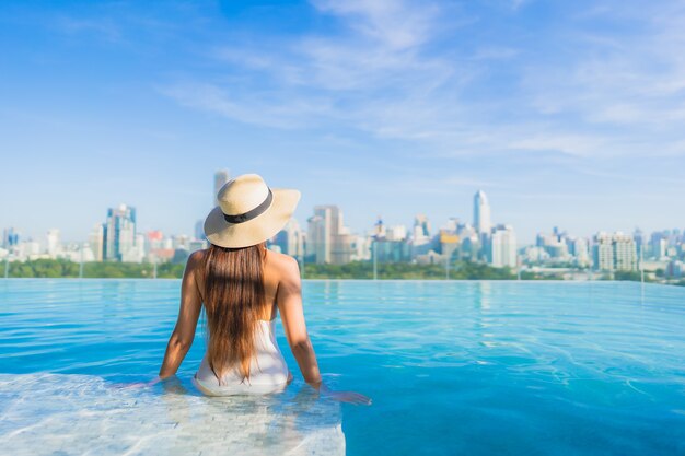 Bella giovane donna asiatica del ritratto che si rilassa intorno alla piscina all'aperto con la vista della città