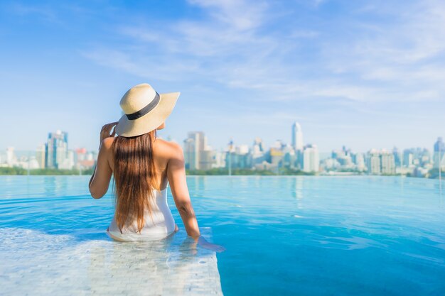 Bella giovane donna asiatica del ritratto che si rilassa intorno alla piscina all'aperto con la vista della città