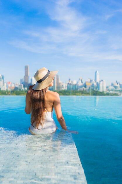 Bella giovane donna asiatica del ritratto che si rilassa intorno alla piscina all'aperto con la vista della città