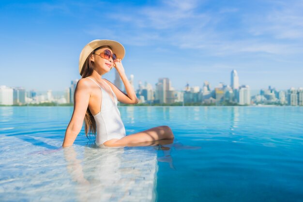 Bella giovane donna asiatica del ritratto che si rilassa intorno alla piscina all'aperto con la vista della città