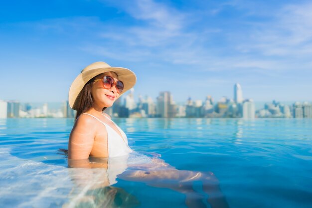 Bella giovane donna asiatica del ritratto che si rilassa intorno alla piscina all'aperto con la vista della città