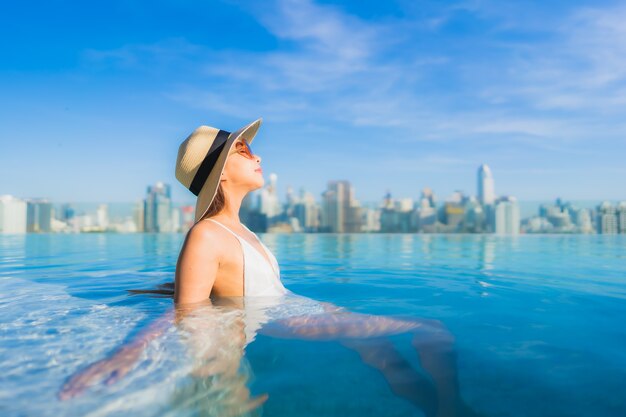 Bella giovane donna asiatica del ritratto che si rilassa intorno alla piscina all'aperto con la vista della città