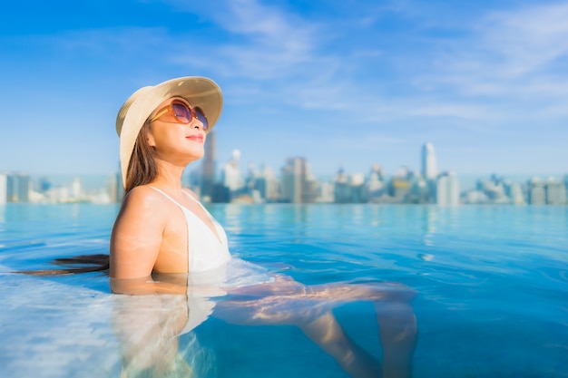 Bella giovane donna asiatica del ritratto che si rilassa intorno alla piscina all'aperto con la vista della città