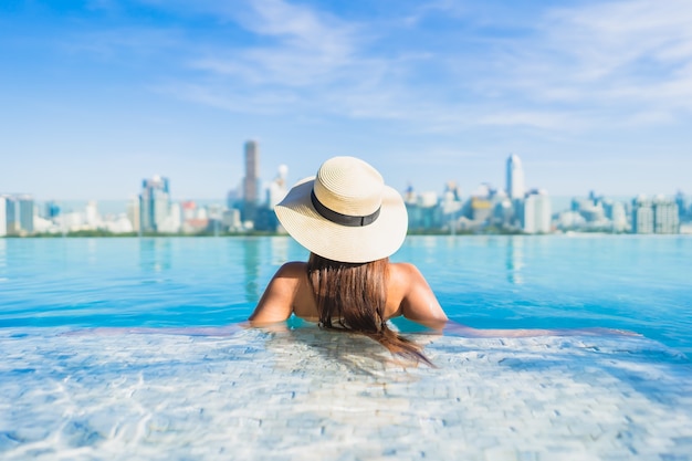 Bella giovane donna asiatica del ritratto che si rilassa intorno alla piscina all'aperto con la vista della città