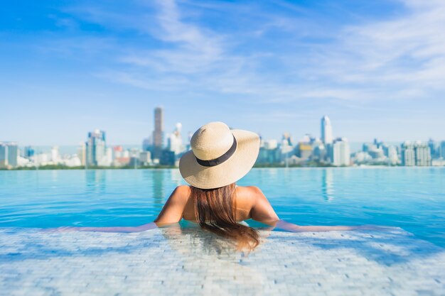 Bella giovane donna asiatica del ritratto che si rilassa intorno alla piscina all'aperto con la vista della città