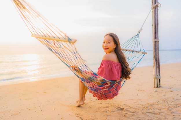 Bella giovane donna asiatica del ritratto che si siede sull&#39;amaca con il mare ed il oce felici vicini della spiaggia di sorriso