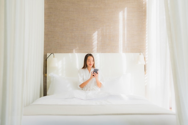 Bella giovane donna asiatica del ritratto che utilizza telefono cellulare astuto sul letto nella camera da letto