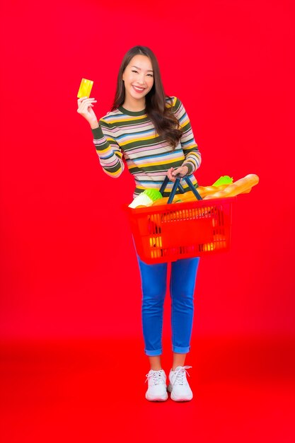 Bella giovane donna asiatica del ritratto con il cestino della drogheria dal supermercato sulla parete isolata rossa