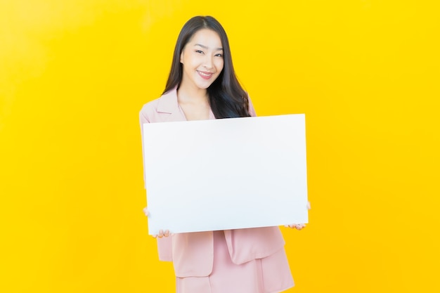 Bella giovane donna asiatica del ritratto con il tabellone per le affissioni bianco vuoto sulla parete gialla