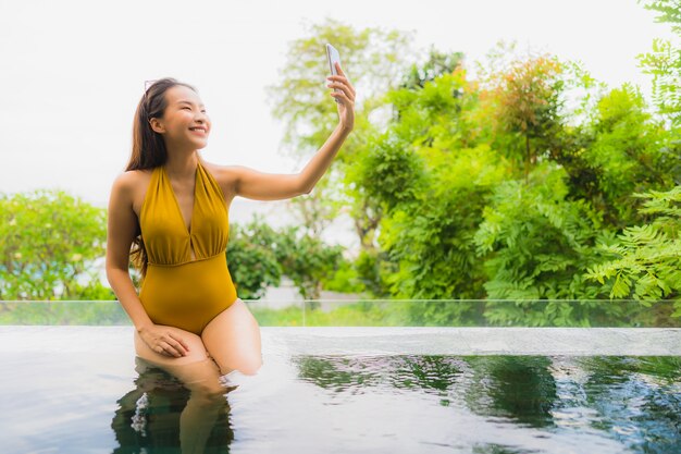 Bella giovane donna asiatica del ritratto con il telefono cellulare o il telefono cellulare intorno alla piscina alla località di soggiorno dell&#39;hotel