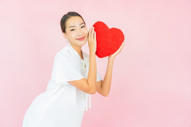 Bella giovane donna asiatica del ritratto con la forma del cuscino del cuore sulla parete rosa