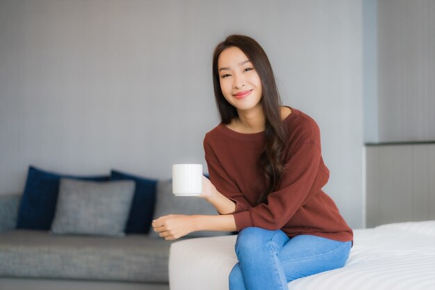 Bella giovane donna asiatica del ritratto con la tazza di caffè sul letto nell'interno della camera da letto