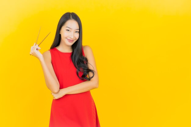 Bella giovane donna asiatica del ritratto con le bacchette pronte da mangiare sulla parete gialla