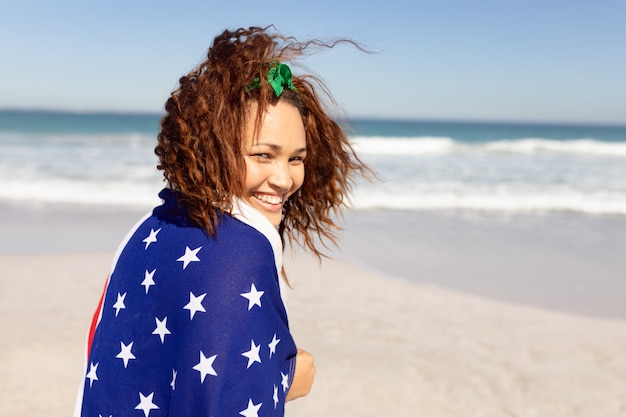 Bella giovane donna avvolta in bandiera americana che guarda l'obbiettivo sulla spiaggia al sole