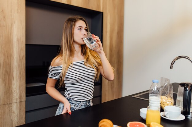 Bella giovane donna bionda che beve acqua in cucina con cibo sano sul tavolo