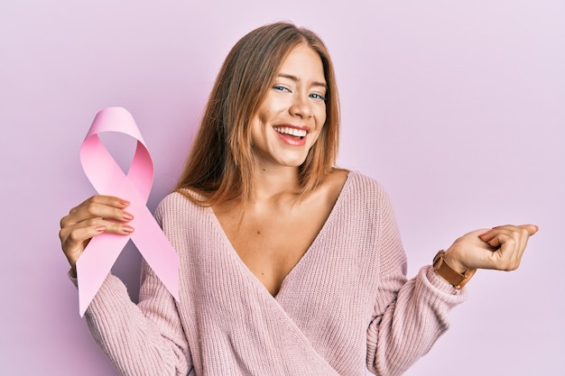 Bella giovane donna bionda che tiene il nastro rosa del cancro urlando orgoglioso, celebrando la vittoria e il successo molto eccitato con il braccio alzato