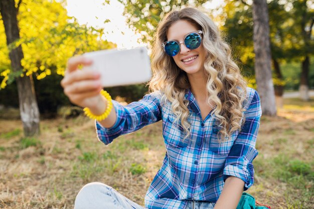 Bella giovane donna bionda sorridente attraente alla moda che si siede nel parco, stile casual estivo