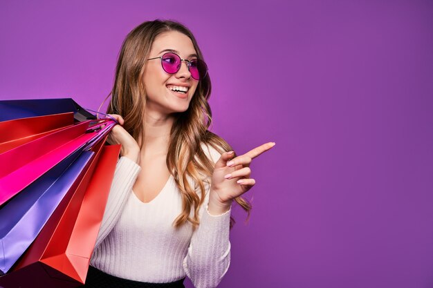 Bella giovane donna bionda sorridente che indica in occhiali da sole che tengono borse della spesa e carta di credito su una parete rosa