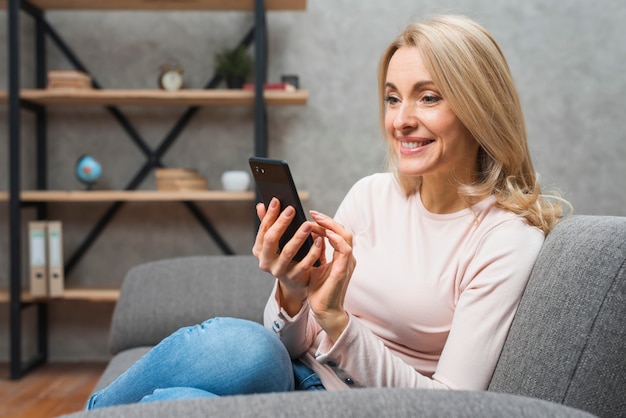 Bella giovane donna bionda sorridente che si siede sul sofà facendo uso dello Smart Phone