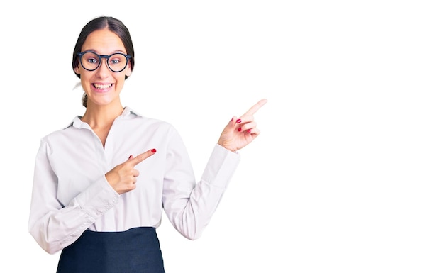 Bella giovane donna castana che indossa il grembiule da cameriera professionale sorridendo e guardando la telecamera che punta con due mani e le dita di lato.