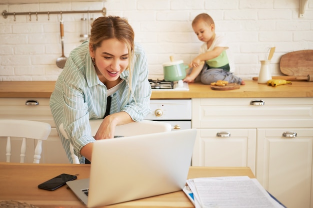 Bella giovane donna che cerca di lavorare utilizzando laptop e babysitter suo figlio neonato. Bambino sveglio che si siede sul bancone della cucina, giocando con la casseruola, sua madre che scrive sul computer portatile in primo piano