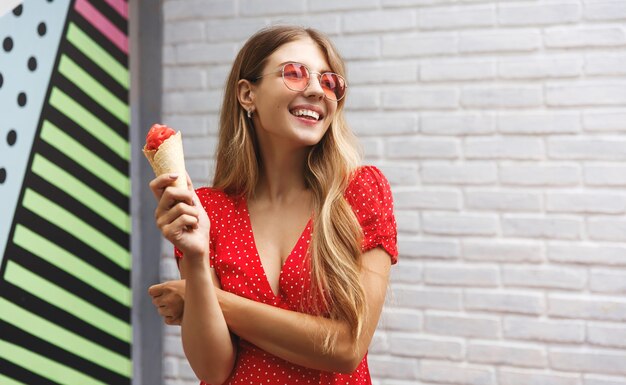 Bella giovane donna che gode del cibo di strada dolce e sorridente, esplora la città durante le vacanze estive, indossando occhiali da sole con vestito