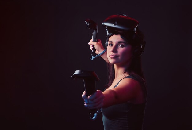Bella giovane donna che indossa l'auricolare per realtà virtuale e tiene i joystick, posando una fotocamera ar. Isolato su sfondo scuro.