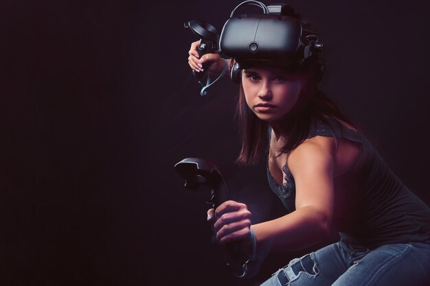 Bella giovane donna che indossa l'auricolare per realtà virtuale e tiene i joystick, posando una fotocamera ar. Isolato su sfondo scuro.