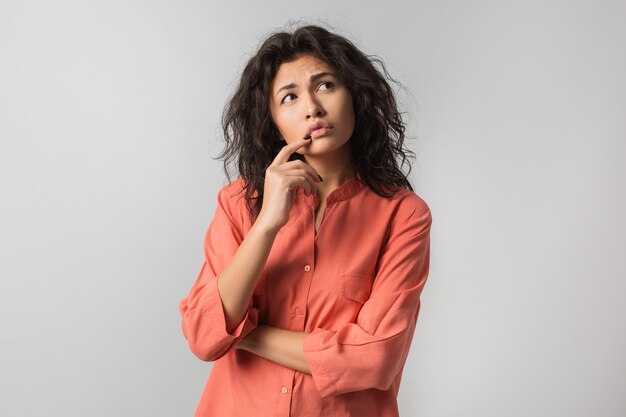 Bella giovane donna che pensa al problema, emozione confusa, isolato, indossa una maglietta arancione, stile hipster, capelli ricci, tenendo il dito sulle labbra, guardando in alto, avendo un'idea