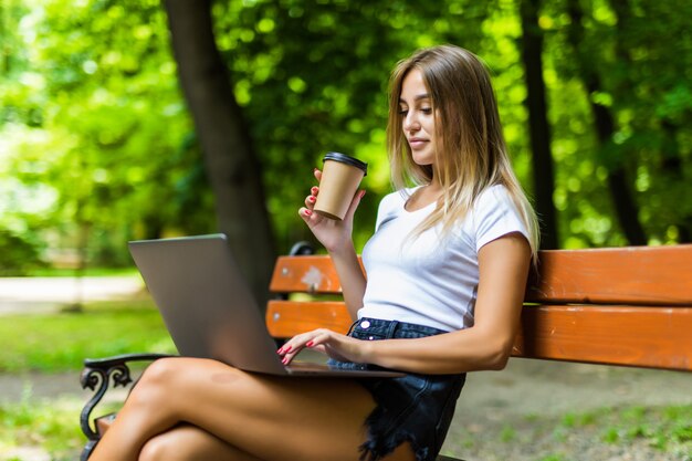 Bella giovane donna che per mezzo del computer portatile mentre sedendosi su un banco, bevendo la tazza di caffè asportabile